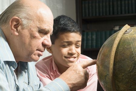Grandfather and grandson are looking at a globe together. Grandfather is pointing at a place on the globe as the grandson  watches happily. This is a horizontally framed photo.の写真素材