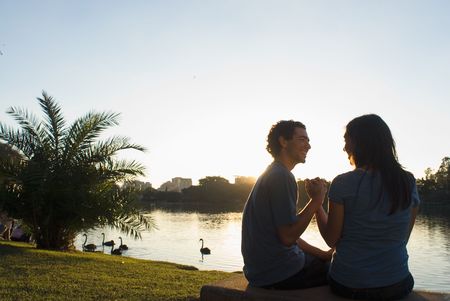 Happy couple clasp hands as they smile at each other. There is a pond with swans and buildings in...