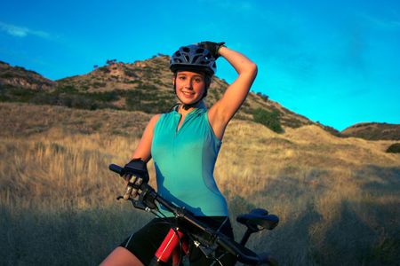 Attractive smiling female mountain-biker, wearing full cycle gear, straddling a mountain-bike adjusting her helmet.  Horizontally framed shot on with golden grasses and a blue sky in the background.の写真素材
