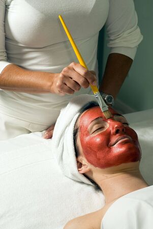 A relaxed smiling woman getting a red facial mask brushed on at a spa.  Vertically framed side-shot.の写真素材