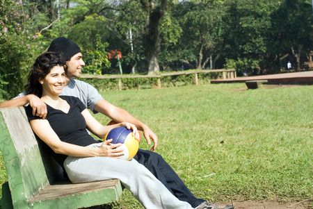 A couple is sitting on a park bench.  They are both smiling and looking away from the camera and each other.  The man has his arm around the woman and the woman is holding a volleyball.  Horizontally framed photo.の写真素材