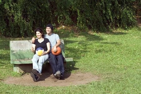 A couple is sitting on a park bench.  They are both smiling and laughing and looking away from the camera and each other.  The man is holding a basketball and the woman is holding a volleyball.  Horizontally framed photo.の写真素材