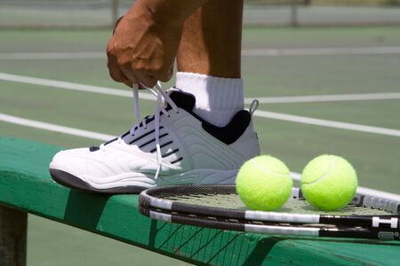 A tennis player tying his shoe on top of a bench.  Also includes view tennis balls, racket, and tennis court.  Horizontally framed shot.の写真素材