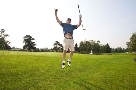 A man is jumping up and down on a golf course.  He is holding a golf club and smiling at the camera.  Horizontally framed shot.の写真素材