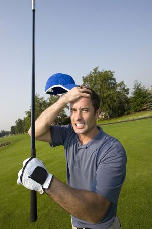A man is grabbing his forehead in anger after missing a shot on a golfcourse.  He is holding a golf club and looking the camera.  Vertically framed shot.の写真素材