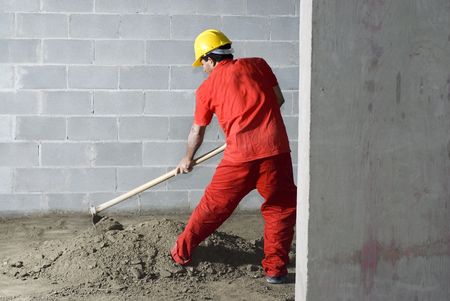 A man is digging through the ground with a hoe on a construction site.  He is looking down at the...