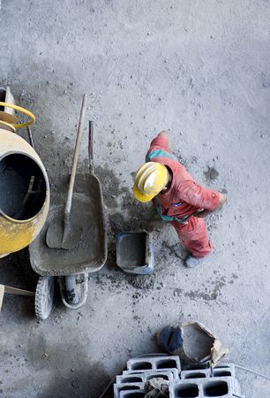 The worker is mixing concrete by cinder blocks in a wheel barrel.  Photo is taking from a vantage point above.  Vertically framed shot.