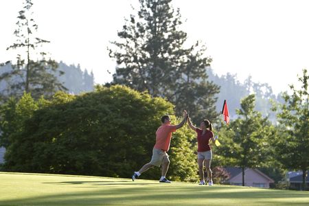 A young couple is playing on a golf course.  The man is holding his golf club and the woman is holding the flag.  They are high fiving and smiling at each other.  Horizontally framed shot.の写真素材