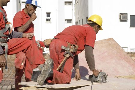 Construction worker cuts board with skill saw. He is wearing a tool belt and two other workers are...