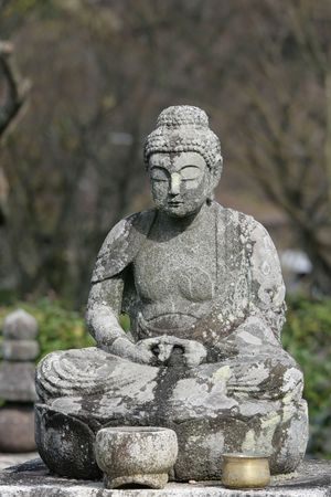 A stone statue of Buddha against a natural setting.  This statue is old and is now covered with lichens.  It's in a meditative pose with a stone and bronze pot in front of him. の写真素材