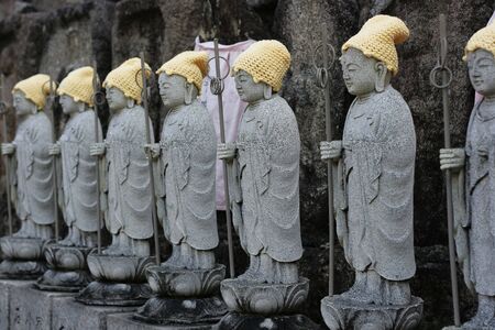 A row of Buddhas with staff in hand.  They have yellow knitted hats and are standing on lotuses. ...