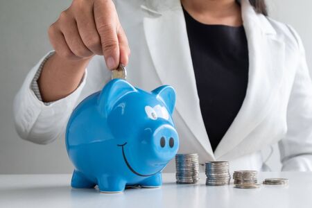 women hand putting coin into blue piggy bank saving money with coins pile.の写真素材