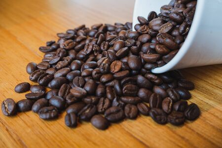 full of coffee beans spilling out white ceramic cup on brown wooden background.の写真素材