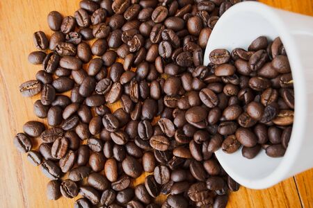 full of coffee beans spilling out white ceramic cup on brown wooden background.の写真素材