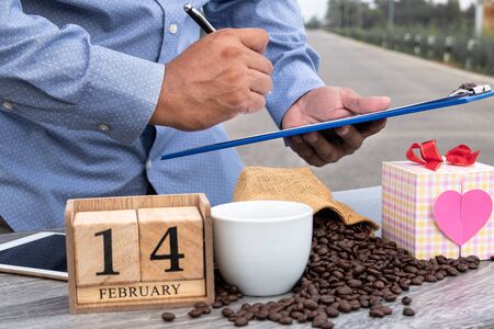 A man uses a laptop and drinks coffee in nature with a heart shaped gift box and wooden calendar....