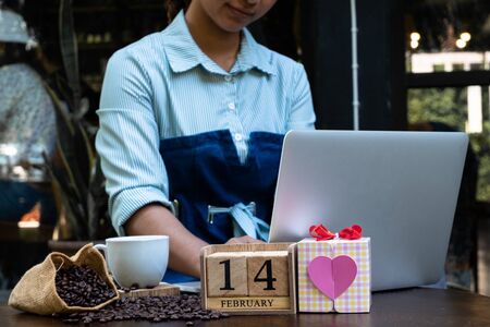 Woman using a laptop in a coffee shop with a gift box with a heart shape and a wooden calendar....