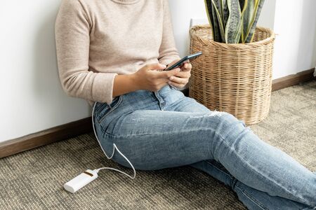 The girl's hand is holding a smart phone and a modern spare battery charger, placed beside to charge the smartphone with a backup battery charger and sitting in the room.の写真素材