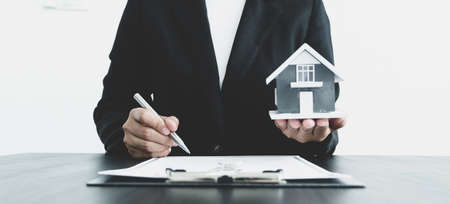 The hand of the real estate agent, holding a pen and drafting an agreement on the home insurance contract documents, along with samples of house models and keys to present to his clients.の写真素材
