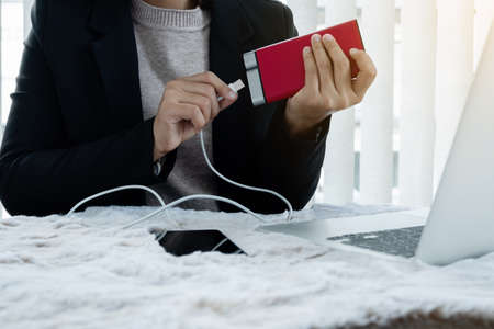 The hand of a young woman holding a smart phone and a modern Power Bank charger. Put it at the desk,...