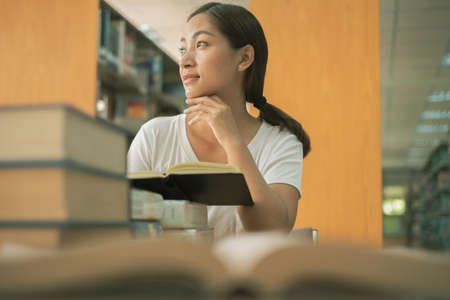 Young Asian women are searching for books and reading from the bookshelves in the college library to...
