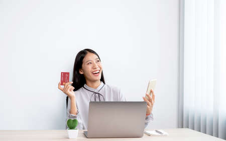 The Asian businesswoman's hand is holding a credit card and using a smartphone and laptop for online...