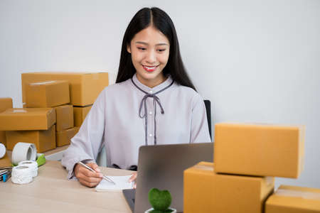 Young Asian woman business owner with many parcel boxes on the table happy online sales job, use your laptop, get an order from customers, take notes, and make arrangements for delivery by post.の写真素材
