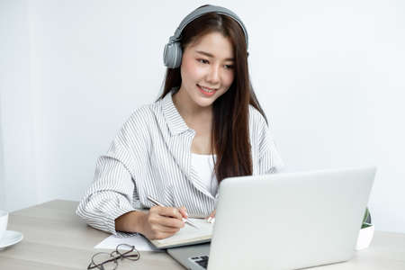 female Asian college student wearing headphones enjoys online tutoring, watching videos and taking notes from digital tablet or doing homework by looking at the screen while sitting at desk at home.の写真素材