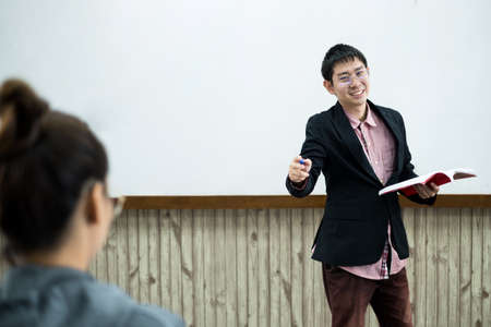 An Asian male teacher in a classroom near the whiteboard is writing and teaching a lesson with a special lecture in a class column. Math teacher explains arithmetic sums to university students.の写真素材