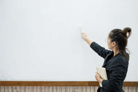 An Asian female teacher in a classroom near the whiteboard is writing and teaching a lesson with a...