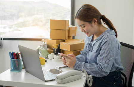 Young Asian woman business owner with many parcel boxes on the table happy online sales job, use the laptop, get an order from customers, take notes, and make arrangements for delivery by post.の写真素材