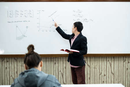 An Asian male teacher in a classroom near the whiteboard is writing and teaching a lesson with a special lecture in a class column. Math teacher explains arithmetic sums to university students.の写真素材