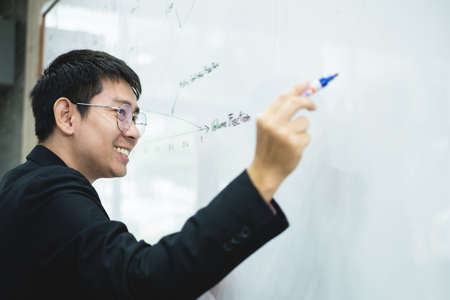 An Asian male teacher in a classroom near the whiteboard is writing and teaching a lesson with a...