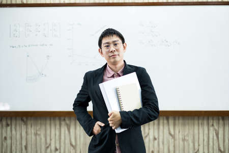 An Asian male teacher in a classroom near the whiteboard is writing and teaching a lesson with a...