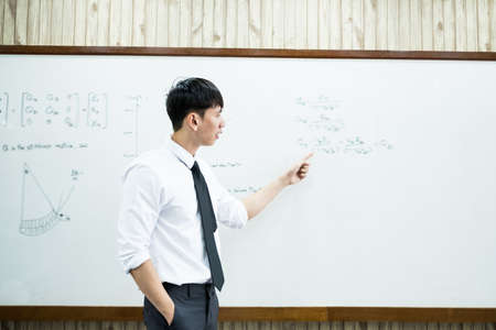 An Asian male teacher in a classroom near the whiteboard is writing and teaching a lesson with a...