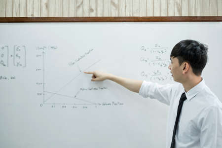 An Asian male teacher in a classroom near the whiteboard is writing and teaching a lesson with a...
