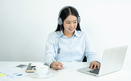 Video call, a young Asian woman wearing headphones, communicates via online video long distance, looking at the digital laptop screen, happily greeted with a friend.の写真素材