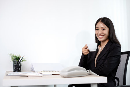 Asian businesswoman working on a laptop with a cheerful and happy smile while working at drink coffee the office.の写真素材