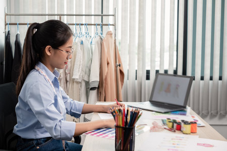 Beautiful young Asian fashion tailor with textile sewing accessories and entrepreneur designer sketches are full of bright colors on the desk with a laptop for creative ideas in the studio.の写真素材