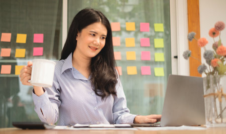 Asian businesswoman smiling happily with a new morning holding a cup of coffee lightly sipping coffee professional woman in size working in the office.の写真素材
