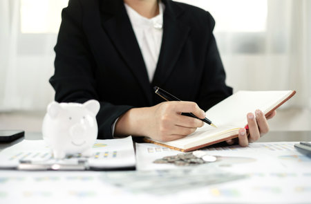 Woman calculating her expenses on a notebook putting coin into piggy bank. Saving money with coins Step into a business that is growing to be successful and save for retirement concepts.の写真素材
