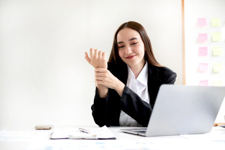 An Asian woman is sitting overtime with her laptop and her wrist aches while working at the office.の写真素材