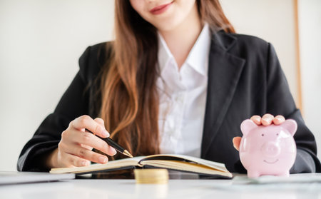 Asian woman calculating her expenses on a notebook putting coin into piggy bank. Saving money with coins Step into a business that is growing to be successful and save for retirement concepts.の写真素材
