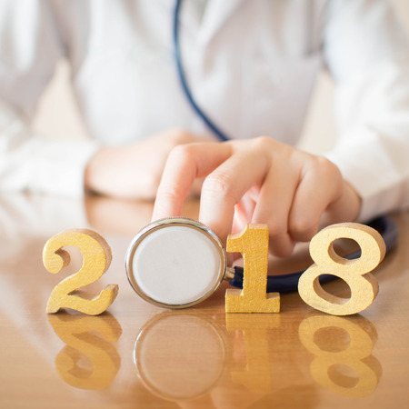 Happy New Year for health care and medical concept. Doctor in white uniform holding stethoscope near gold wooden number 2018 on doctor desk. Idea for new trend in medicine treatment and diagnosis.の写真素材
