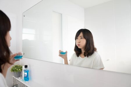 Young asian woman rinsing and gargling mouth with mouthwash after brushing her teeth in bathroom. Oral hygiene routine for freshness breath, prevent plaque and gum disease. Dental health care concept.の写真素材