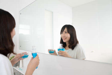 Attractive asian woman smiling and holding mouthwash in front of mirror bathroom. Oral hygiene routine for freshness breath, prevent plaque and gum disease. Dental health care concept.の写真素材