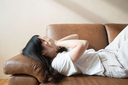 Depressed young asian woman close her face with hands crying and lying on couch at home feels sad, desperate, unhappy, confused and tired. Mental health problem and psychology concept.の写真素材