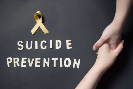 World suicide prevention day - Holding hands for helping and supporting depressed woman with yellow ribbon awareness and SUICIDE PREVENTION wooden word on black background. Mental health care concept.の写真素材
