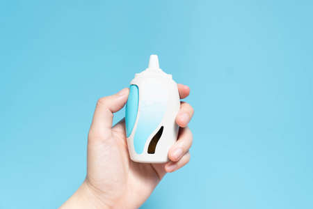 Hand holding steroid nasal spray suspension w/ blank label on light blue background. Medical device...