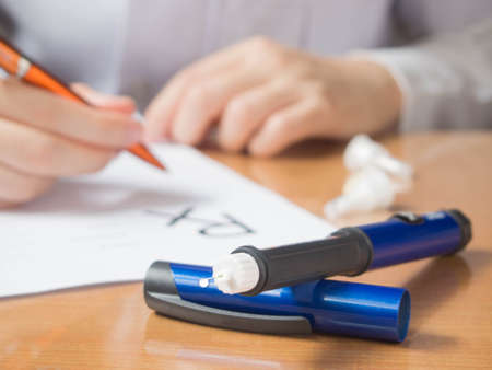 Close up of Insulin pen w/ insulin drop at needle tip on doctor table. Physician writing prescription to diabetes patient at hospital. World diabetes day. Health care medical concept. Selective focus.の写真素材