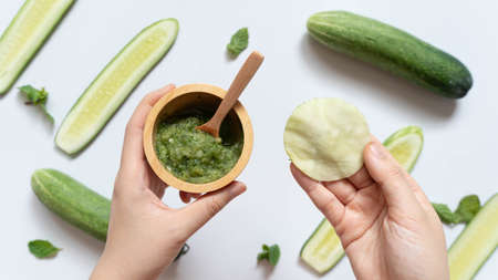 Cucumber mask or makeup remover pads made w/ natural ingredients for healthy skincare. Hands holding DIY eyes/facial mask and a bowl of cucumber mixed w/ mint on white table. Homemade beauty product.の写真素材